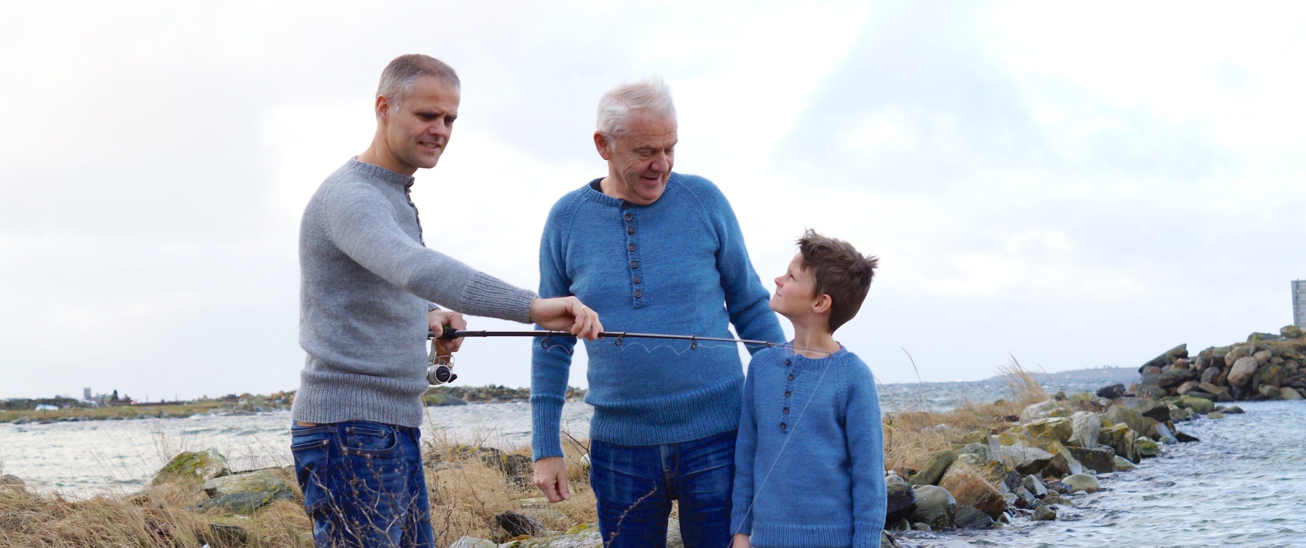 Three generations of men standing by a body of water, possibly fishing.
